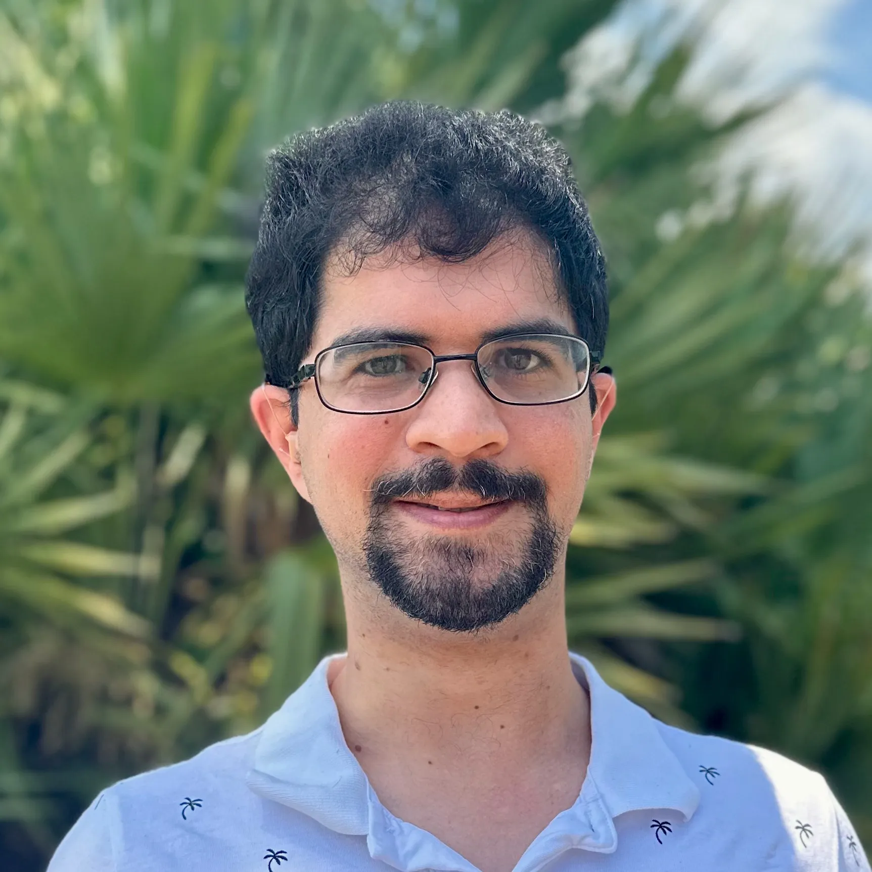 A man with glasses and a white shirt poses in front of a palm tree, enjoying a sunny day outdoors.